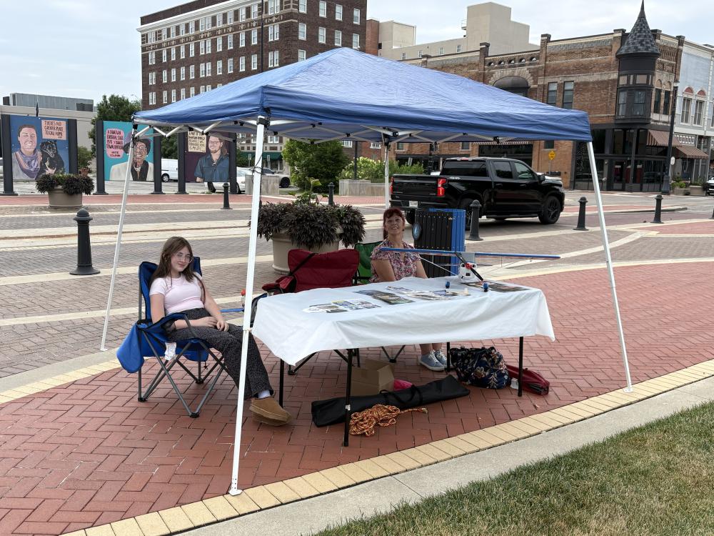 Two people sit under a small blue canopy with a table, on a paved plaza.