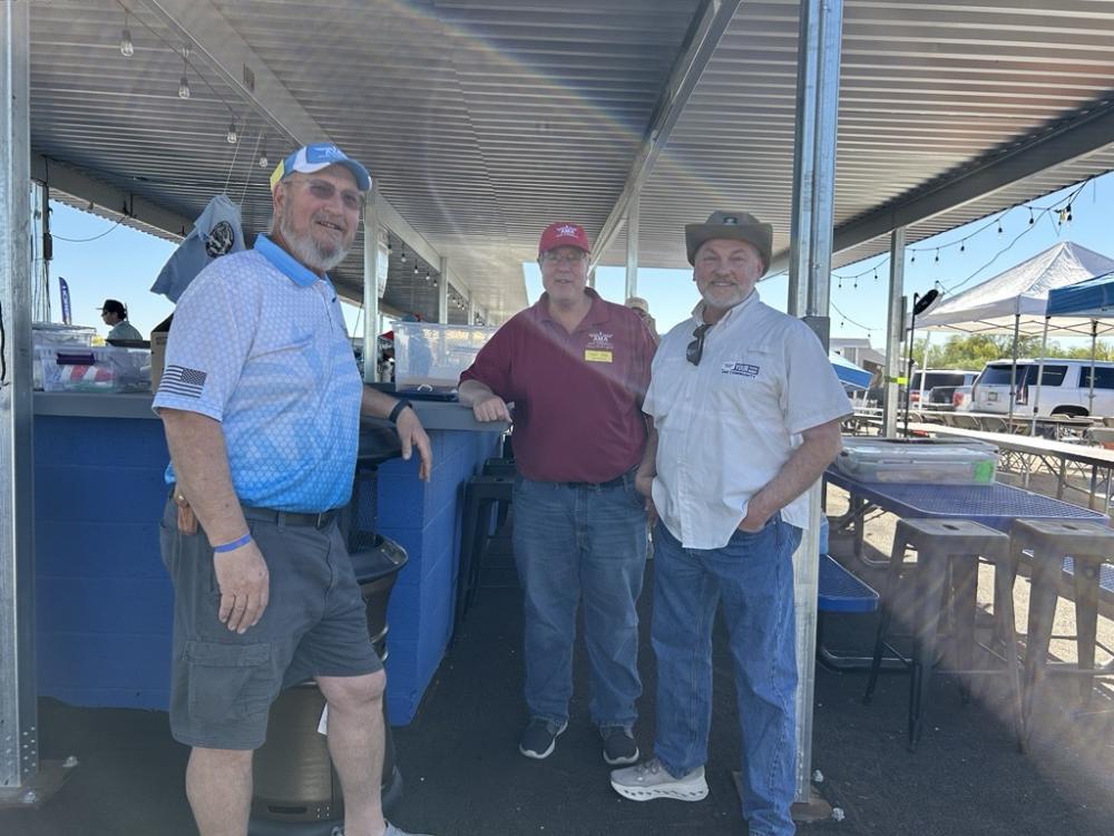 Three men standing together under a pavilion, wearing casual shirts and hats.