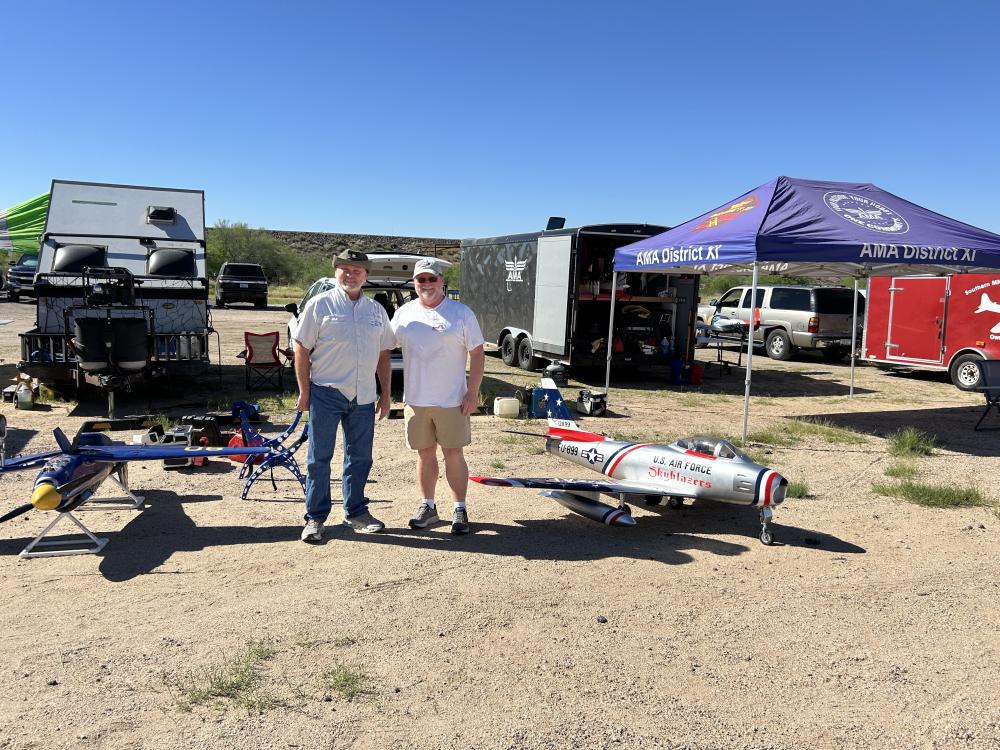 Two people stand by model airplanes at an outdoor event under a clear blue sky.