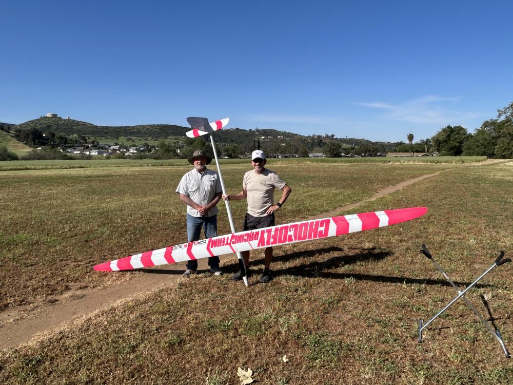 Two people stand on a grassy field with a large pink model glider.
