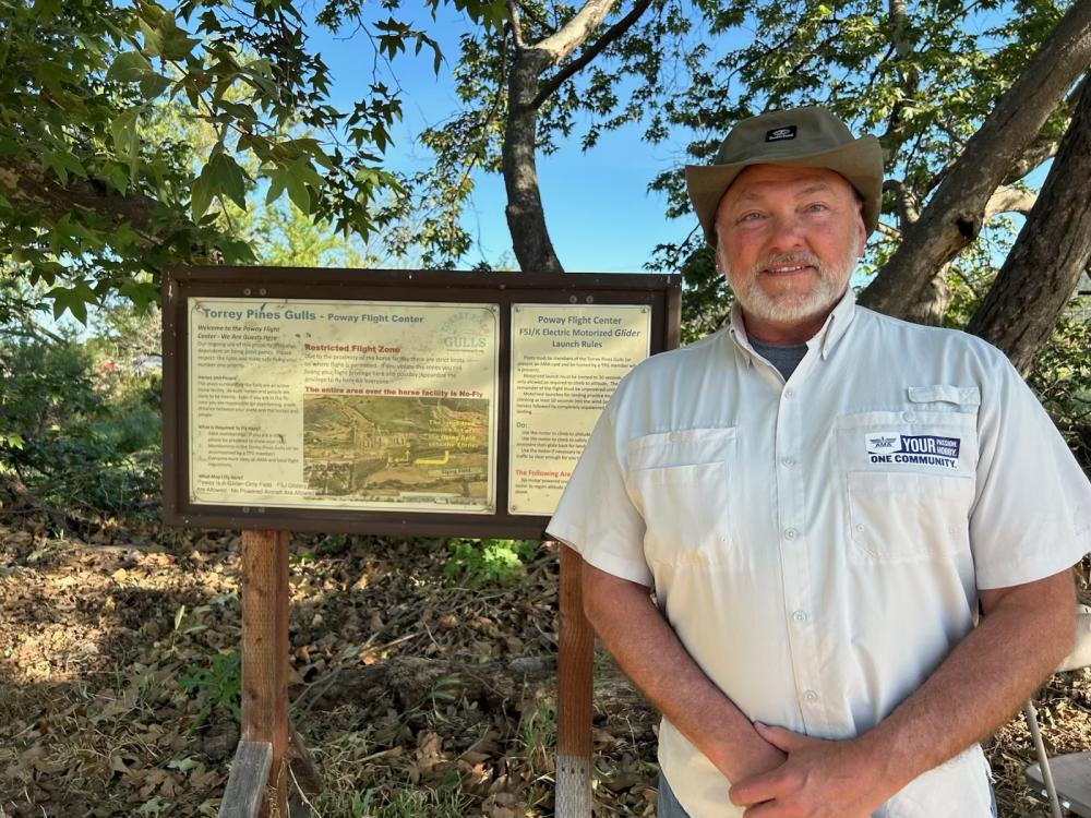 Man in a hat and white shirt standing by an informational board in a wooded area.