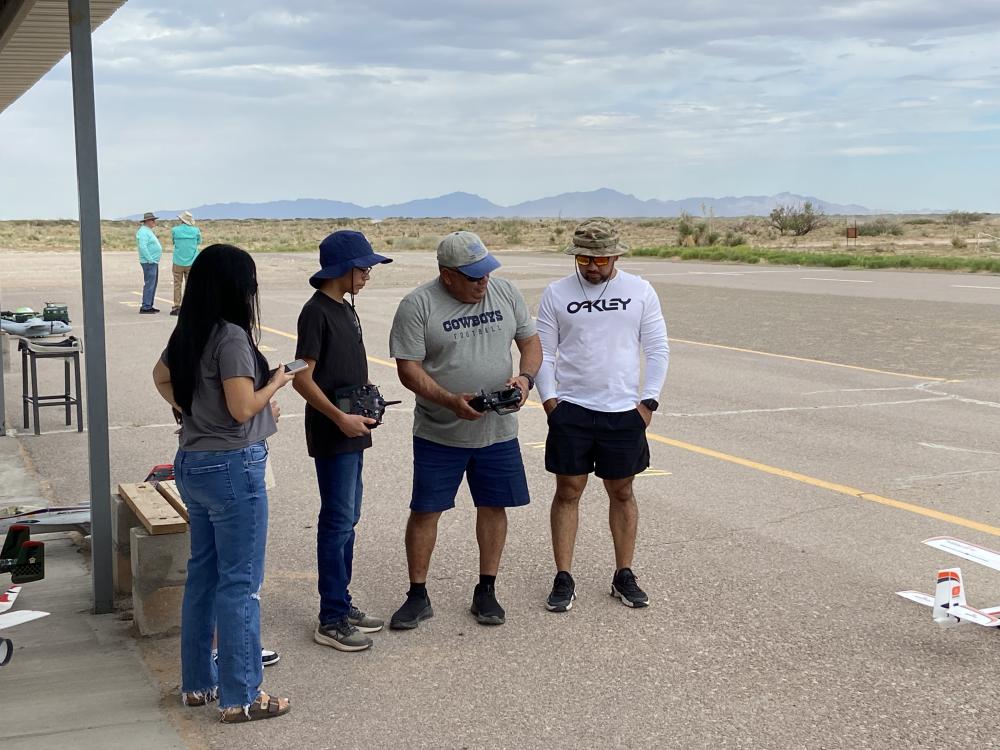 Four people in casual attire at an open airfield, two holding remote controls.
