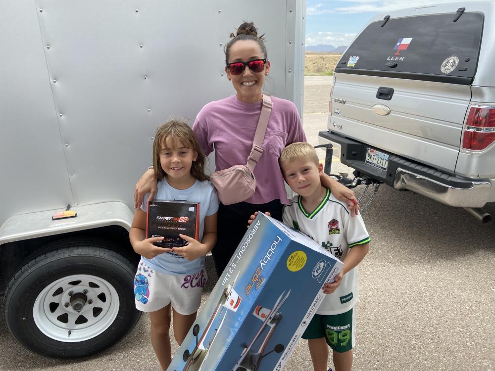 Woman smiling with two children holding boxed toys outdoors near a white trailer.