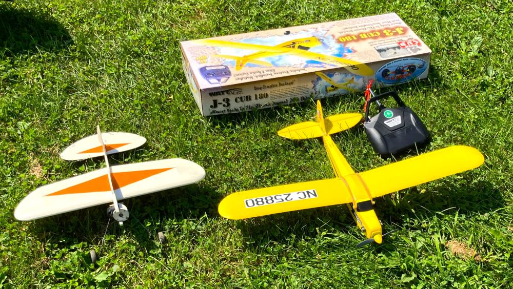Two model airplanes on grass with a cardboard box and a remote control.