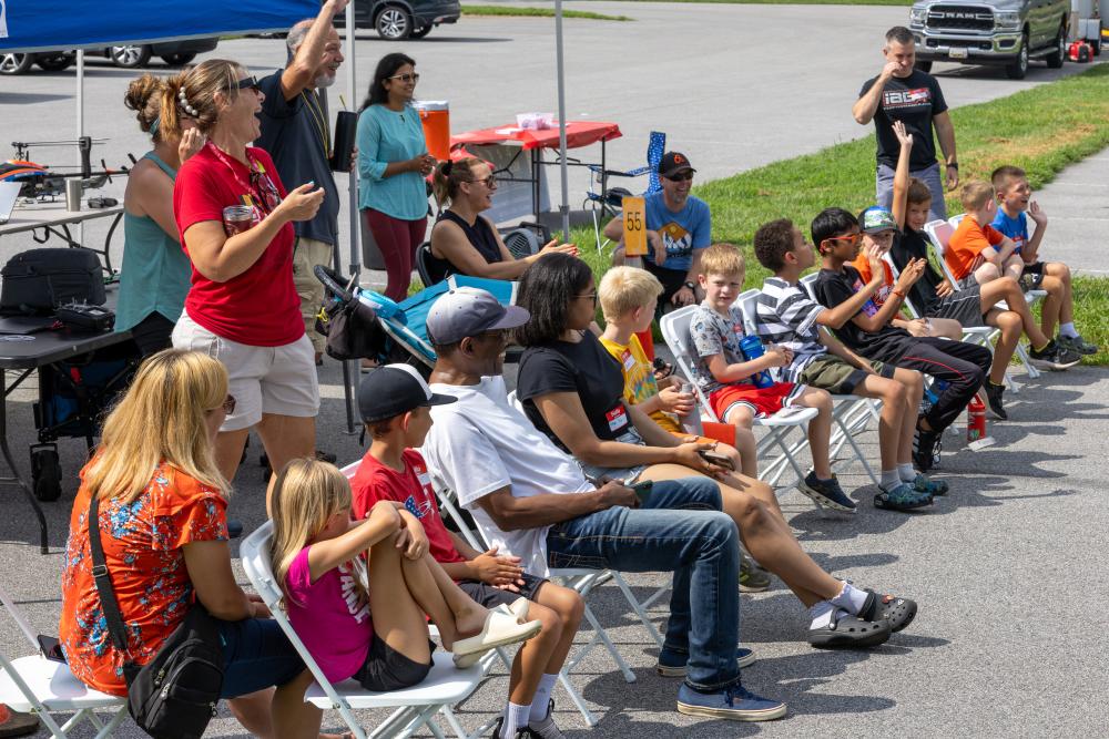 Audience seated outdoors, watching a performance, with kids and adults on folding chairs.