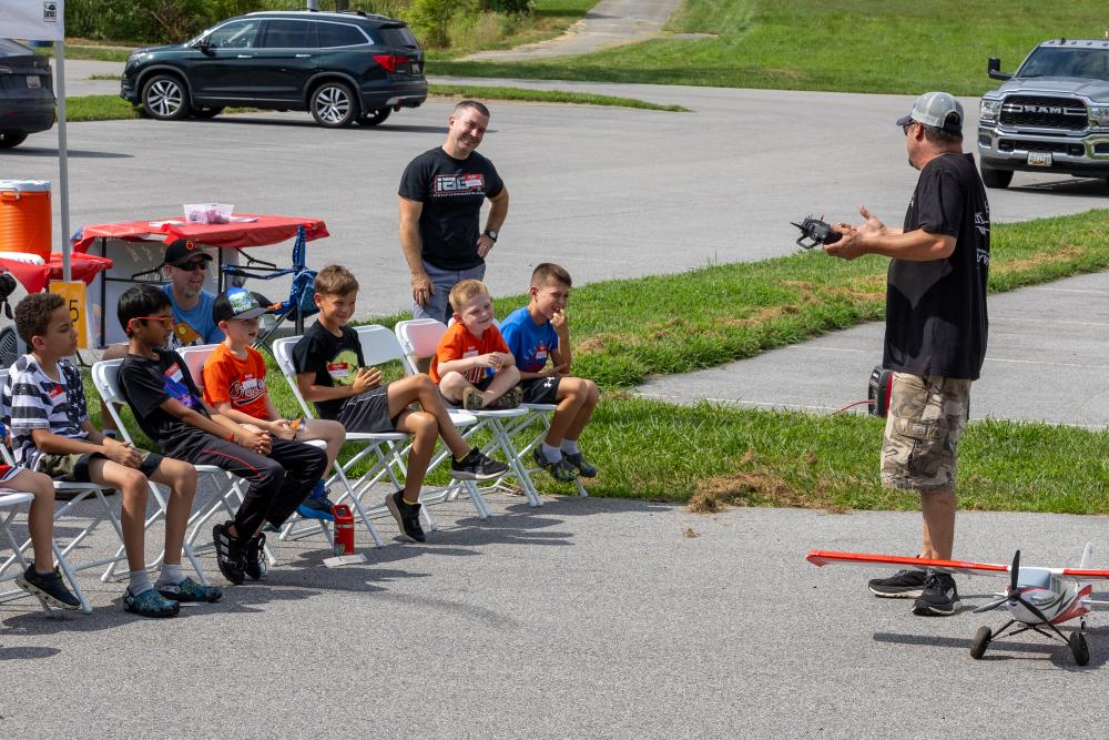 Man demonstrating remote-controlled plane to seated children outdoors.