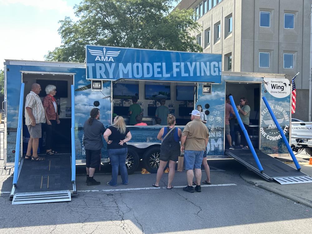 People gathered at a blue trailer promoting model flying in a parking lot.