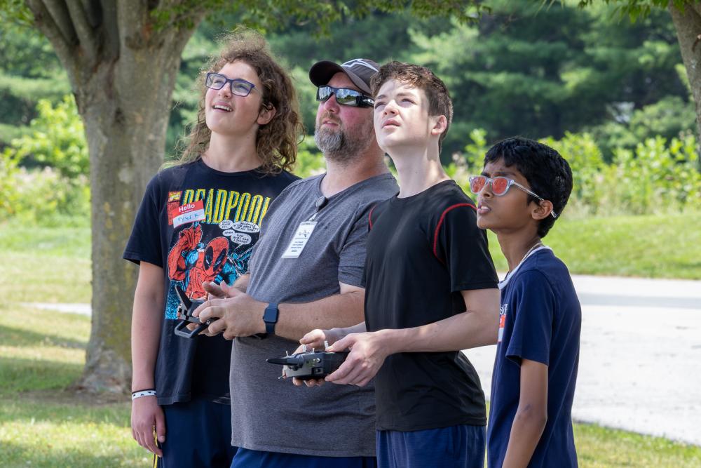 Four people outside, focused on flying drones, trees in the background.