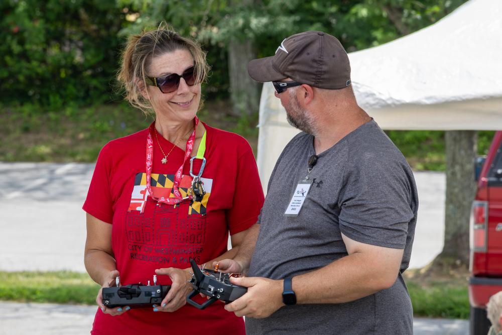 Two people outdoors with remote controllers, talking and smiling.