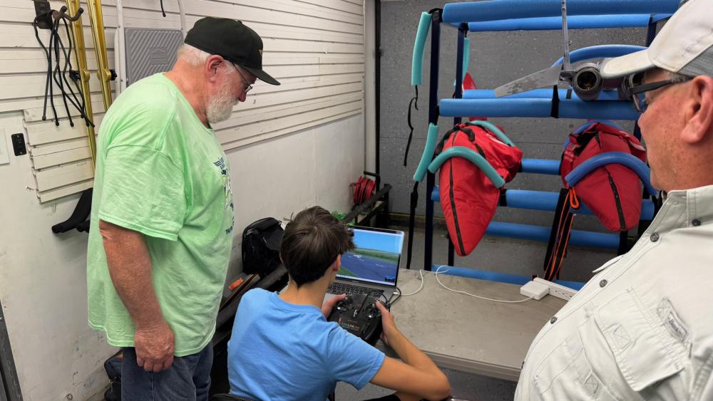 Three men watch a computer screen showing a drone view in a workshop.