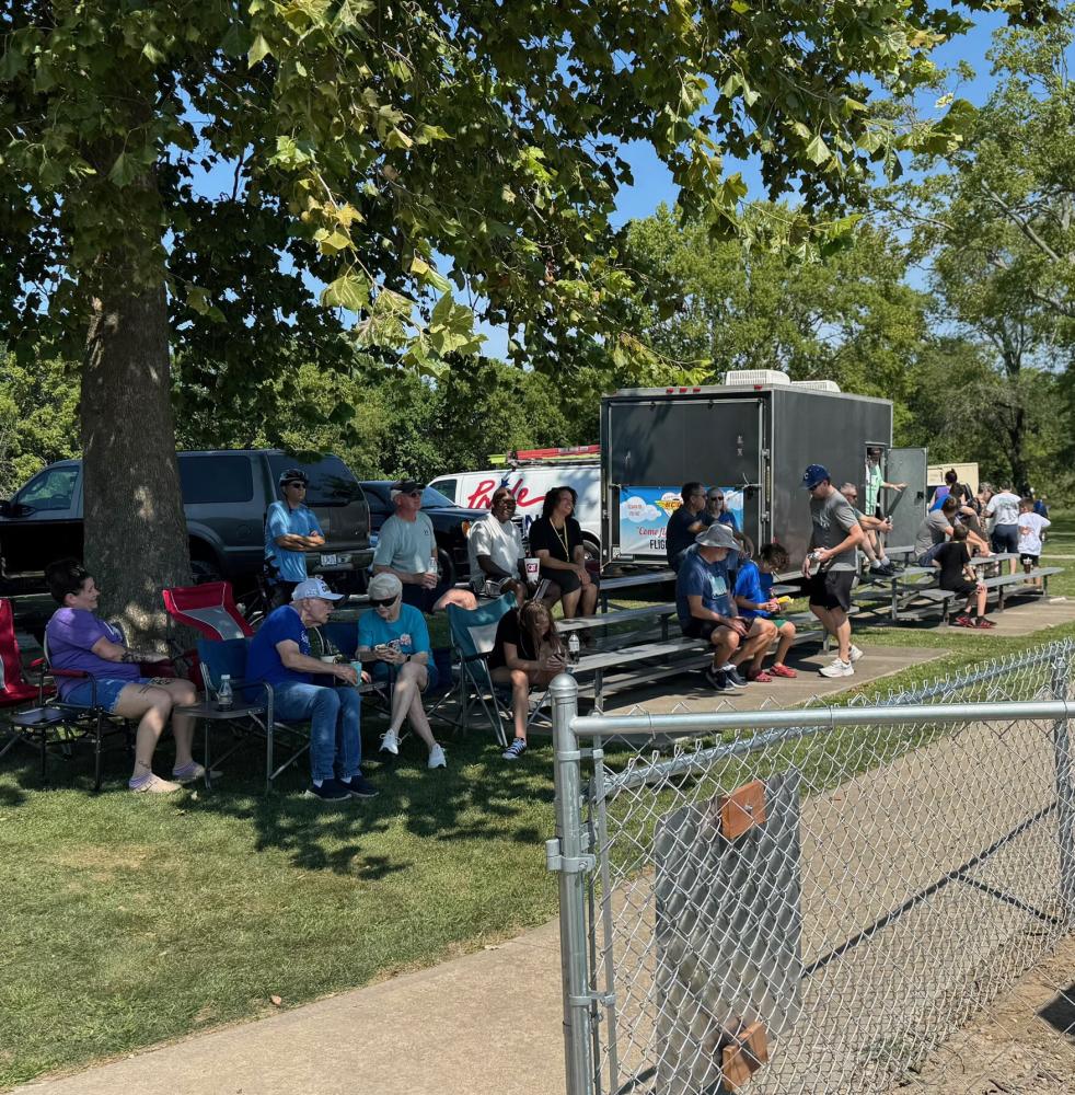 People sitting on bleachers under a tree, watching a game on a sunny day.