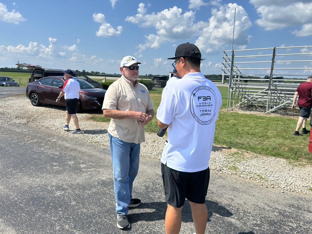 Two men talking outdoors near parked cars and blue sky.