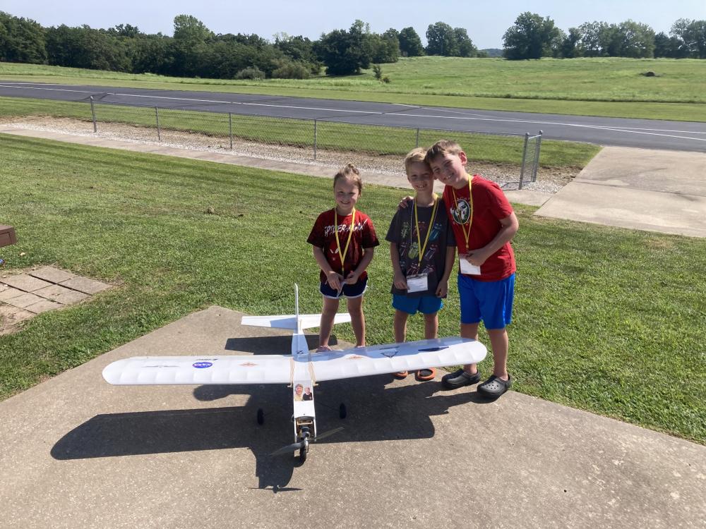 Three children smiling behind a large model airplane on a sunny day.