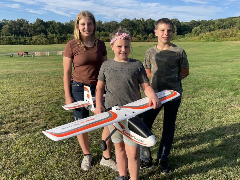 Three kids standing on grass, one holding a remote control plane.