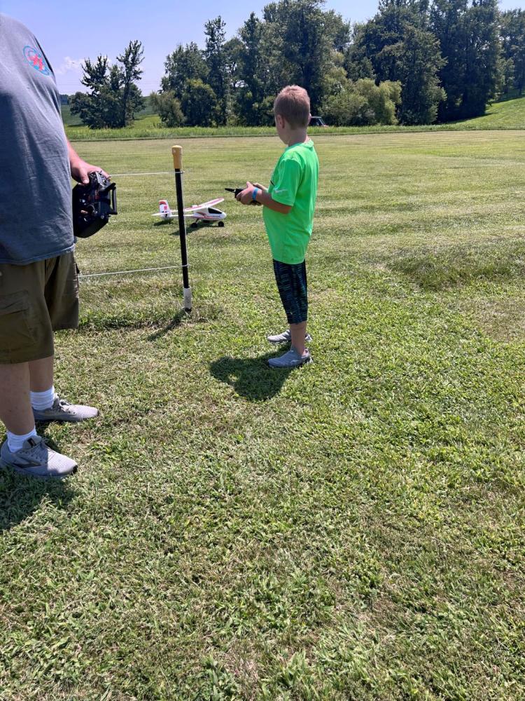 Child in green shirt holding a remote control near a small model airplane on grass.