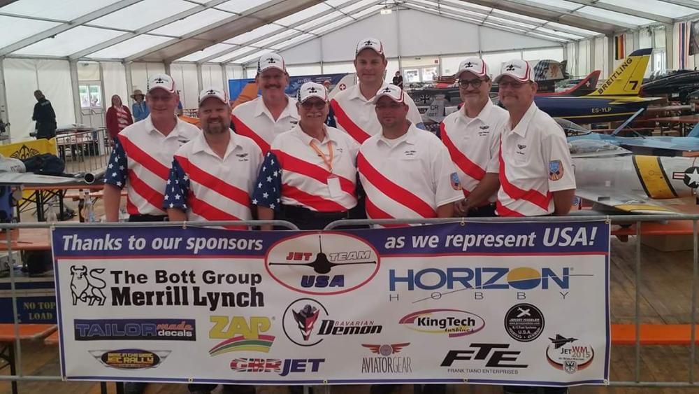 Team in matching shirts posing in front of model planes and sponsor sign in a tent.