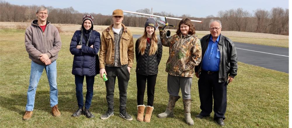 Six people standing on grass, with a model plane in the background.