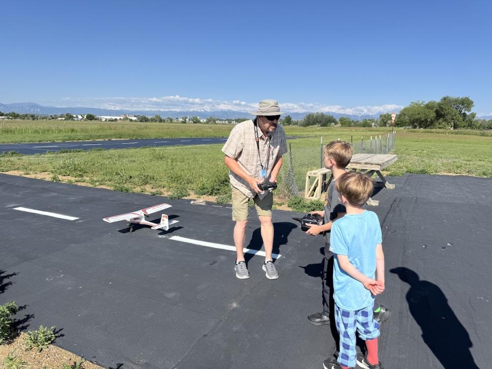 Man instructing two kids with model airplane outdoors on a sunny day.