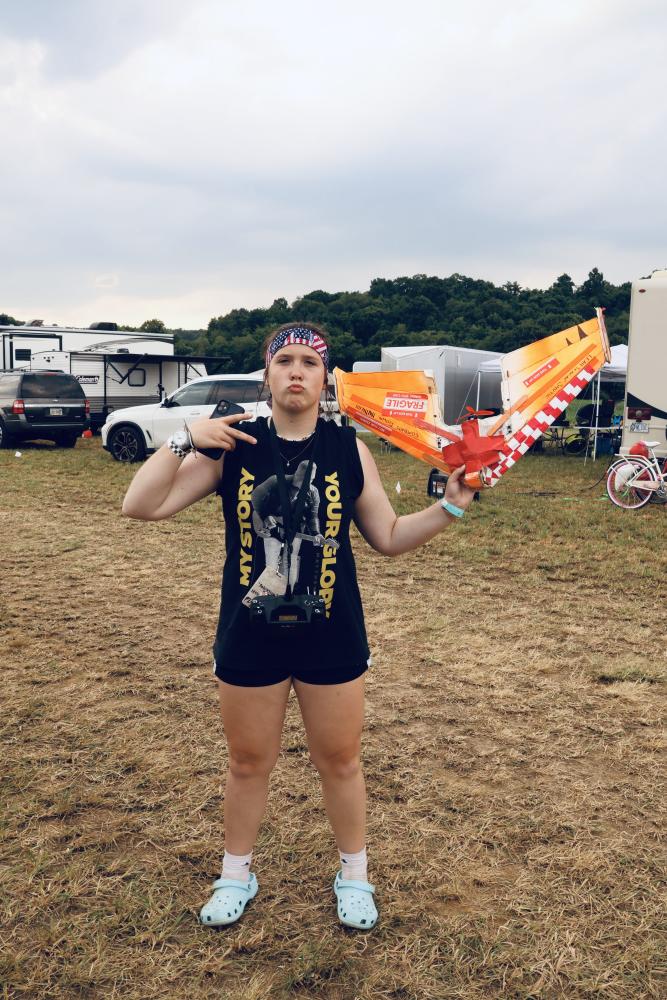 Man outdoors with toy plane, wearing a headband and graphic tank top, showing peace sign.