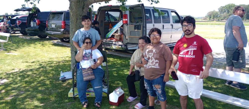 Family poses under a tree at a picnic, with a van and picnic supplies in the background.
