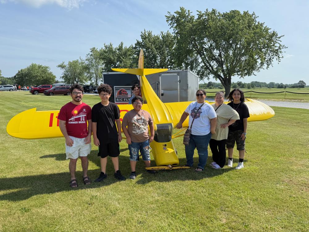 Seven people standing beside a small, yellow vintage airplane on a grassy field.