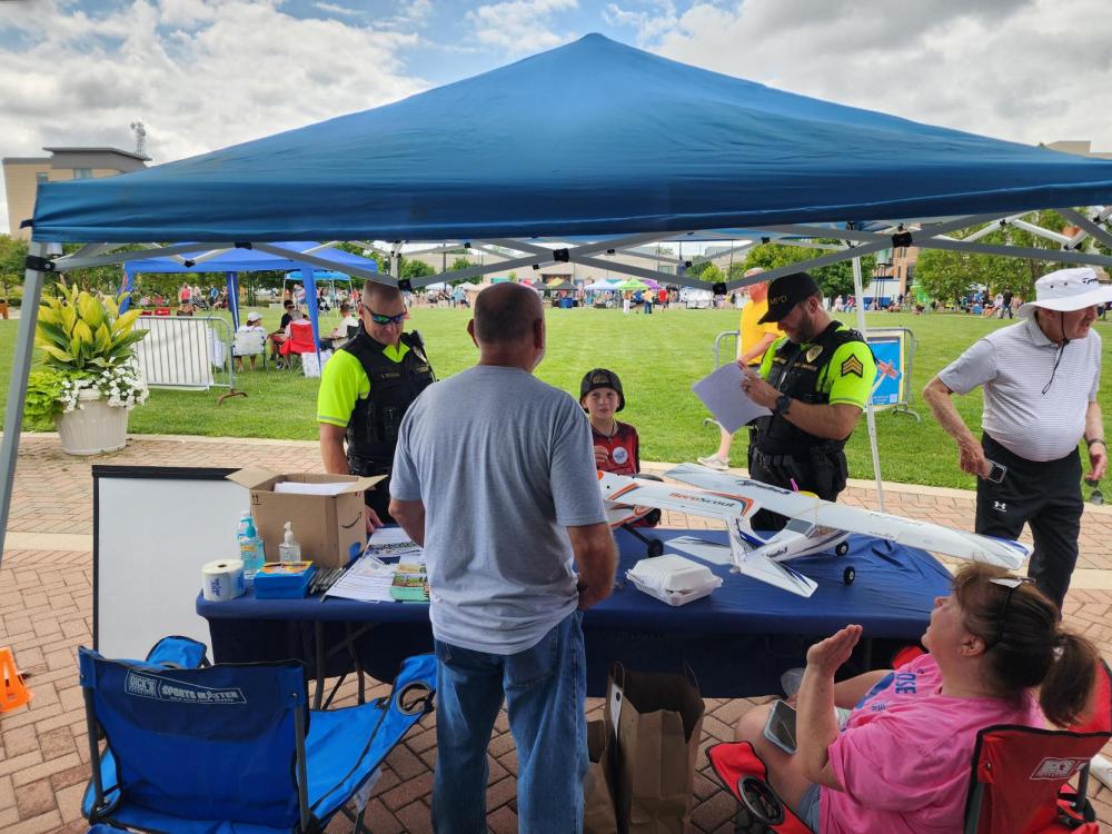 Officers at booth under blue tent, talking with people at outdoor event.