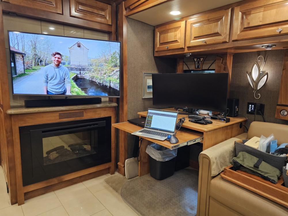 Cozy RV interior workspace with laptop, dual monitors, and a TV showing a man outdoors.