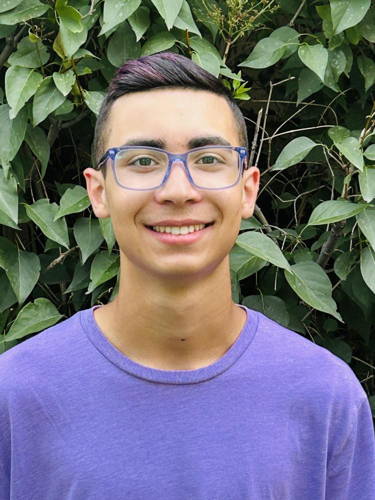 Smiling person with glasses and purple shirt, standing in front of leafy green background.