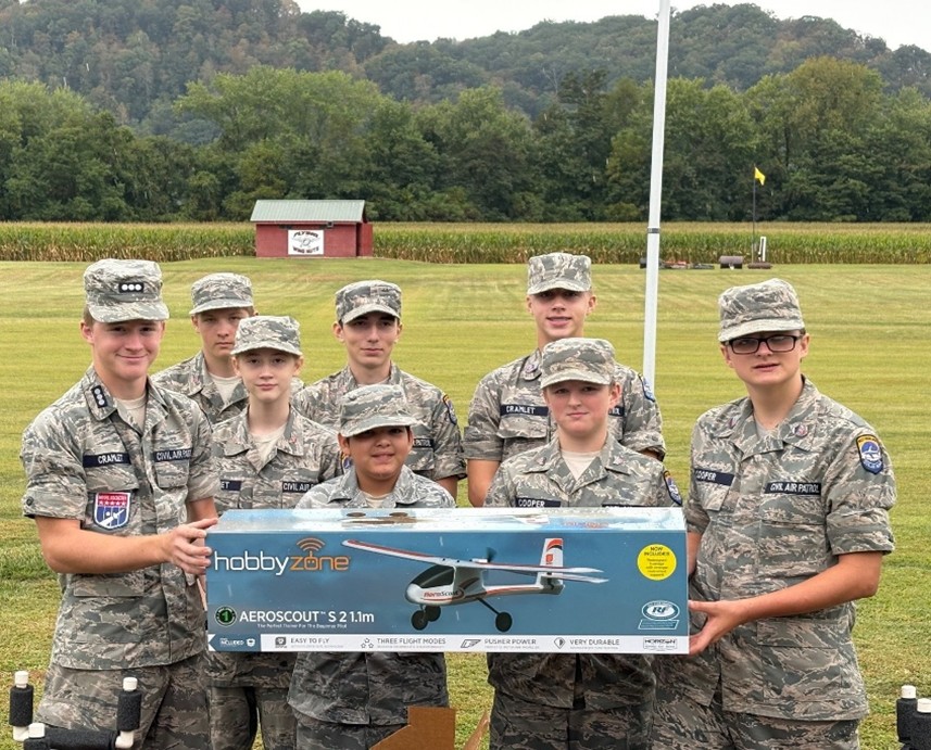 Seven cadets in uniform holding a model airplane box outdoors.