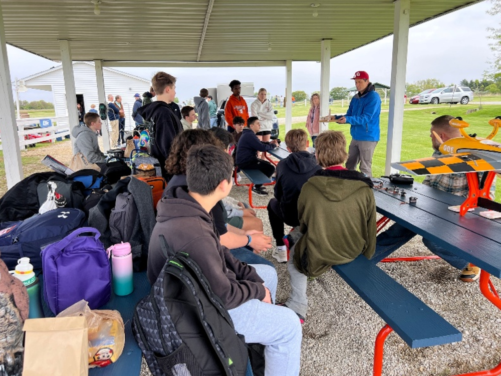 Group of people gathered under a pavilion, some seated, with model planes nearby.