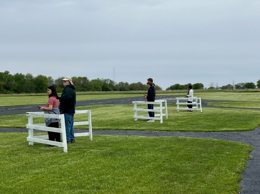 People standing apart at white fences on a grassy field under a cloudy sky.
