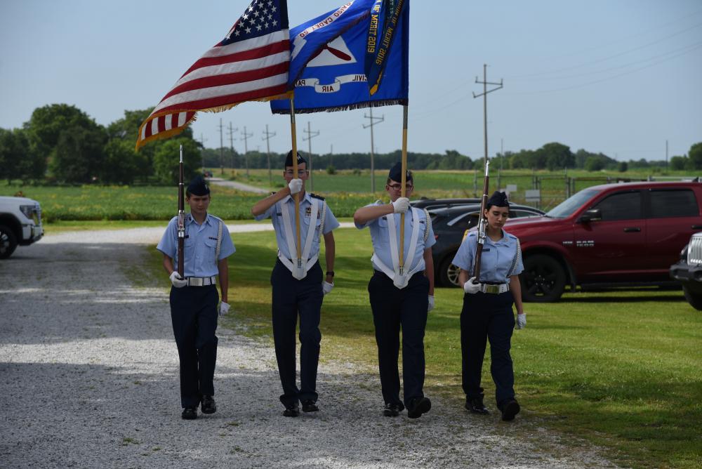Four cadets march, carrying U.S. and Air Force flags, on a gravel path near parked vehicles.