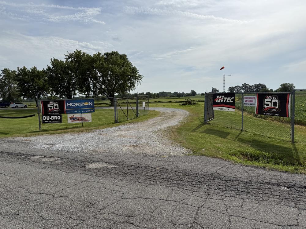 Gravel path leading to a field, lined with sponsor banners on a chain-link fence.