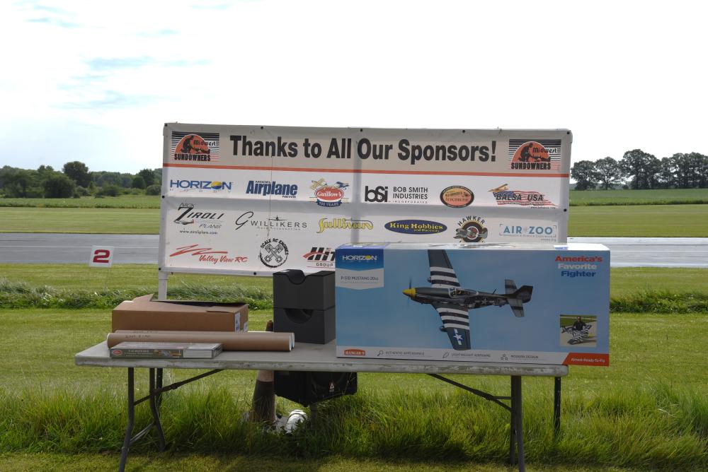 Sign with sponsor logos on grassy field, table with model plane box.