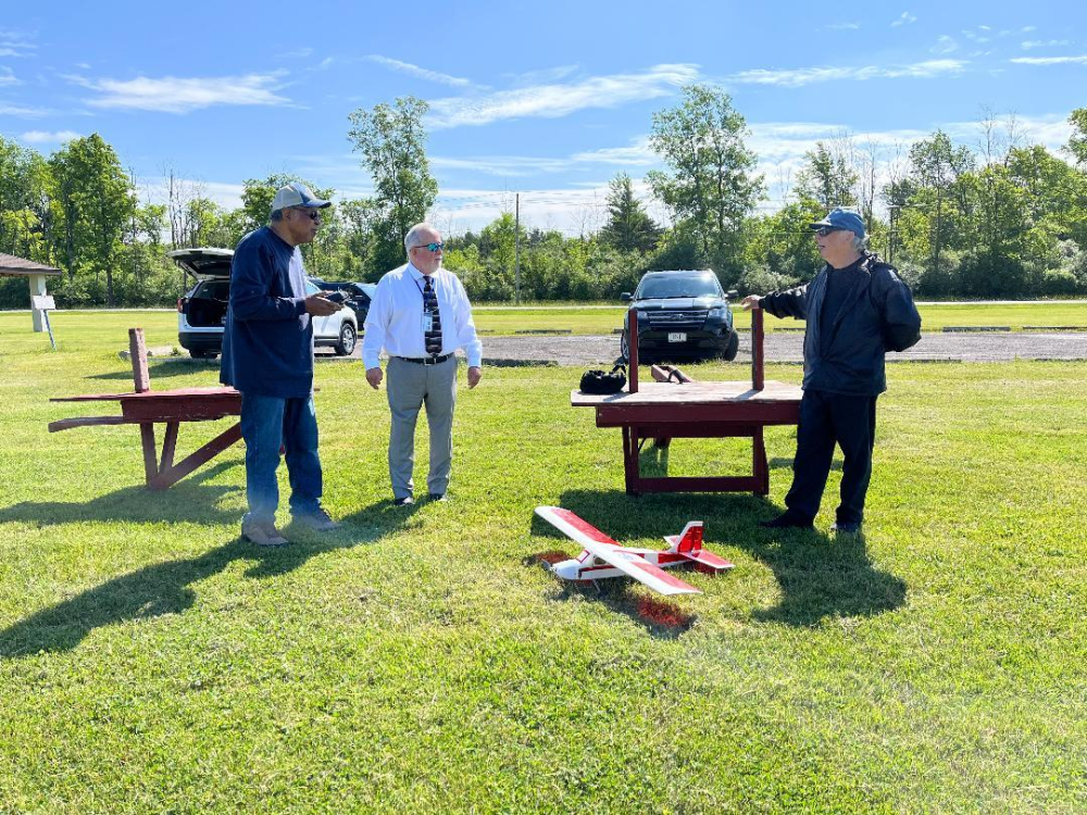 Three men standing on grass near a model airplane, sunny day with cars and trees in background.