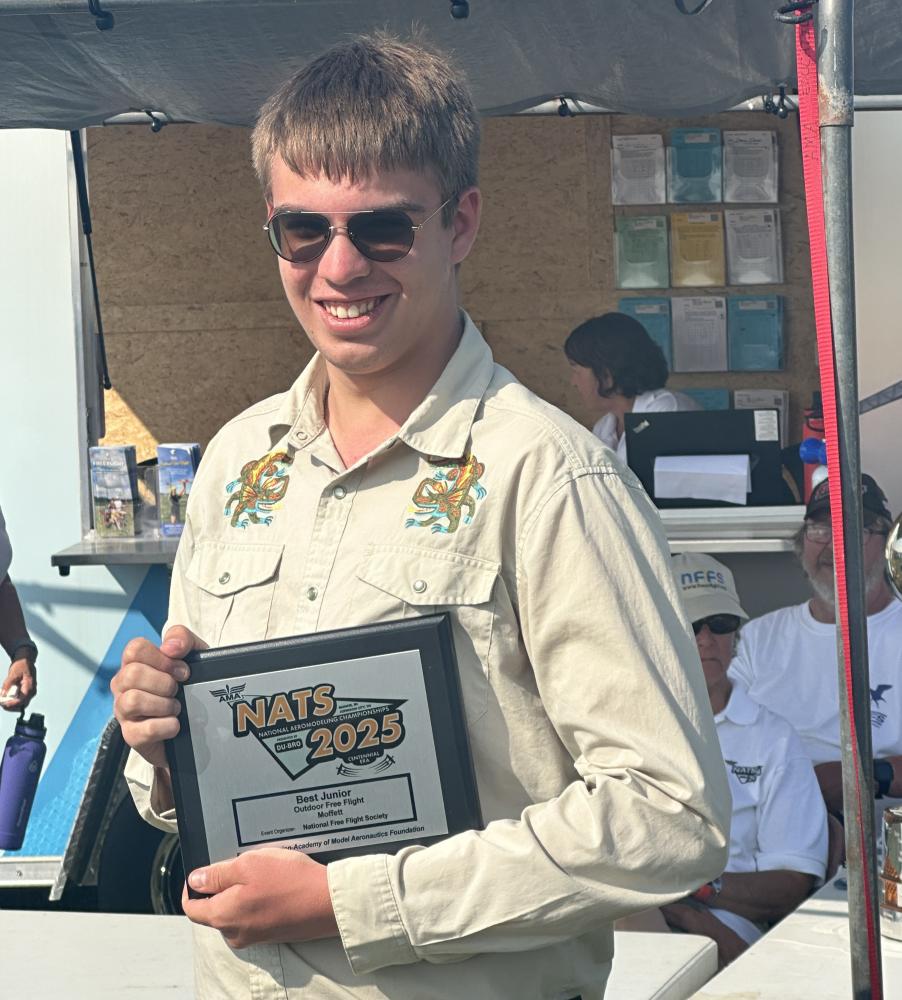 Young man holding a framed award, smiling outdoors in a casual setting.