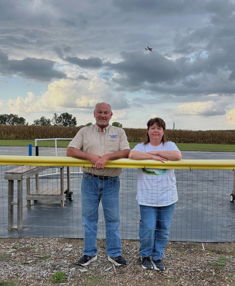 Two people smiling by a fence with a cloudy sky and airplane overhead.