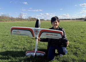 Child kneeling on grass with remote-controlled airplane and controller, smiling.