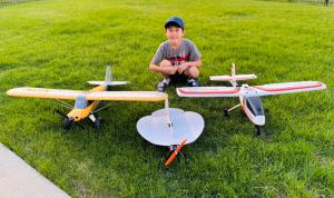 Boy kneeling on grass with three model airplanes.