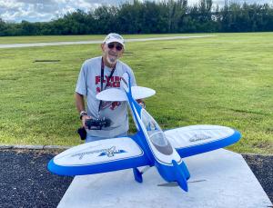 Man holding a remote control beside a large blue model airplane on a grassy field.