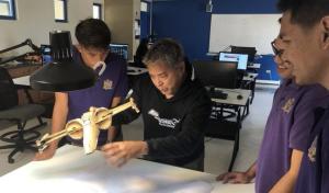 Instructor demonstrating drone parts to three students in a classroom.