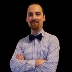 Man in checked shirt and bow tie, arms crossed, against a black background.