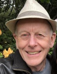 Smiling man in a hat, outdoors, surrounded by greenery and yellow flowers.
