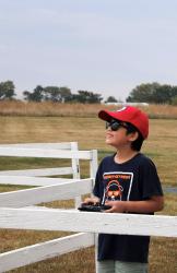 Child in red cap and sunglasses, smiling, holds a remote control, standing by a white fence outdoors.