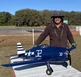 Man smiling with a large blue model airplane outdoors on a sunny day.