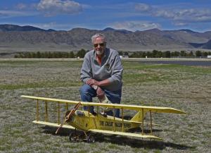 Man kneeling next to a yellow model biplane on a grassy field with mountains in the background.