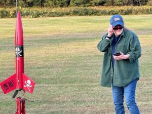 Rocket enthusiast adjusting cap beside a red model rocket on a grassy field.