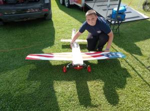 Boy kneeling next to a model airplane with red, white, and blue design on grass.