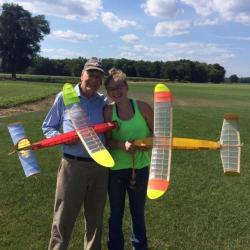 Elderly man and woman smiling, holding colorful model airplanes on a grassy field.
