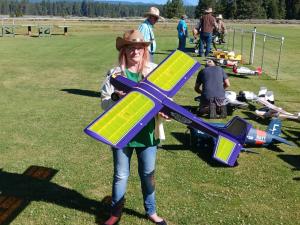 Woman holding large purple and yellow model airplane on grassy field.